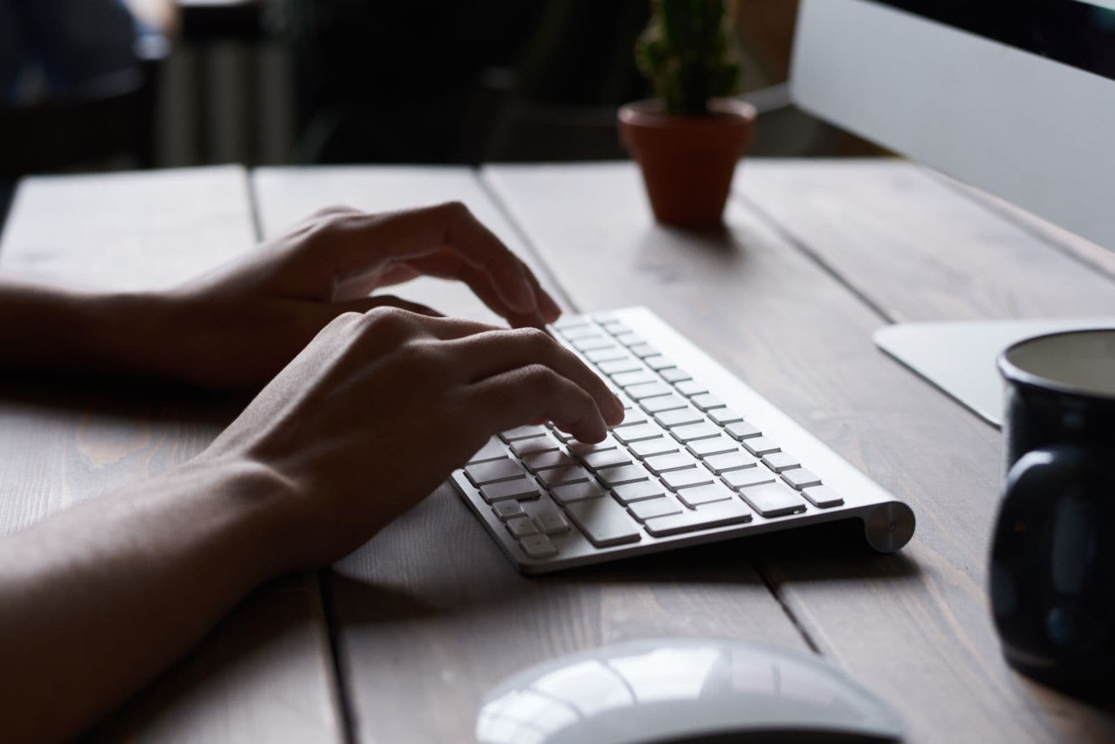 Two hands angled inward to type on a compact white keyboard on a wooden desk, with a mug and a mouse nearby