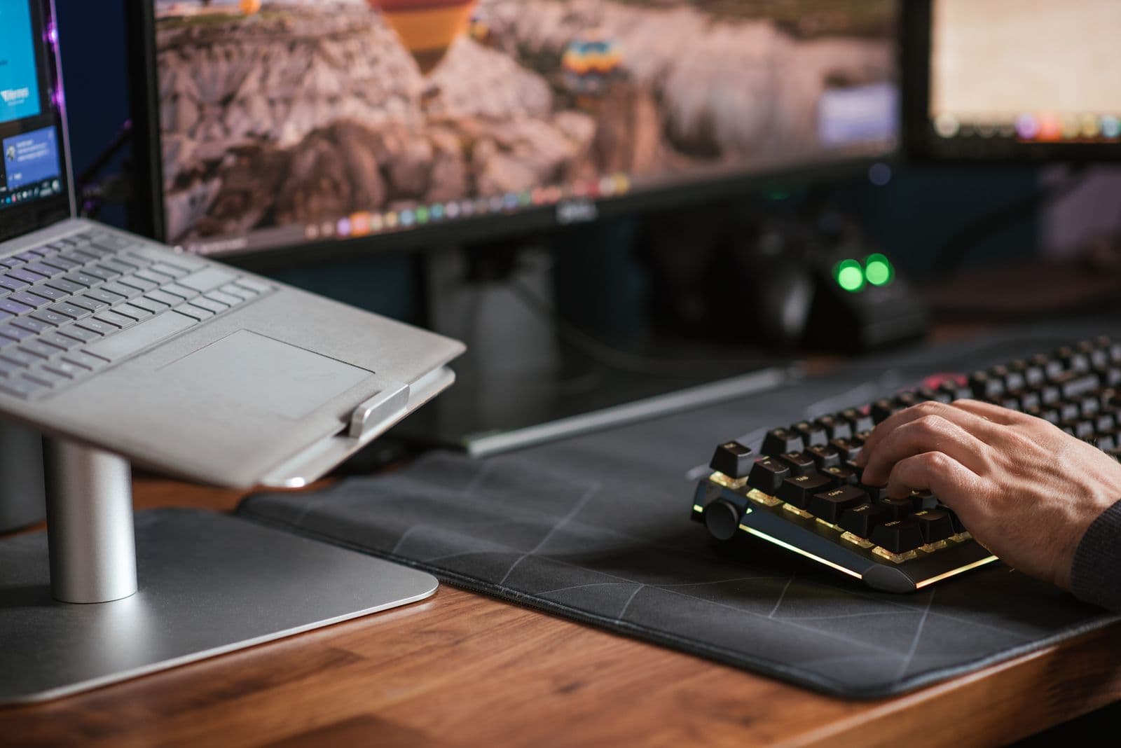 A hand typing on a keyboard resting on a wooden desktop beside a laptop on a stand
