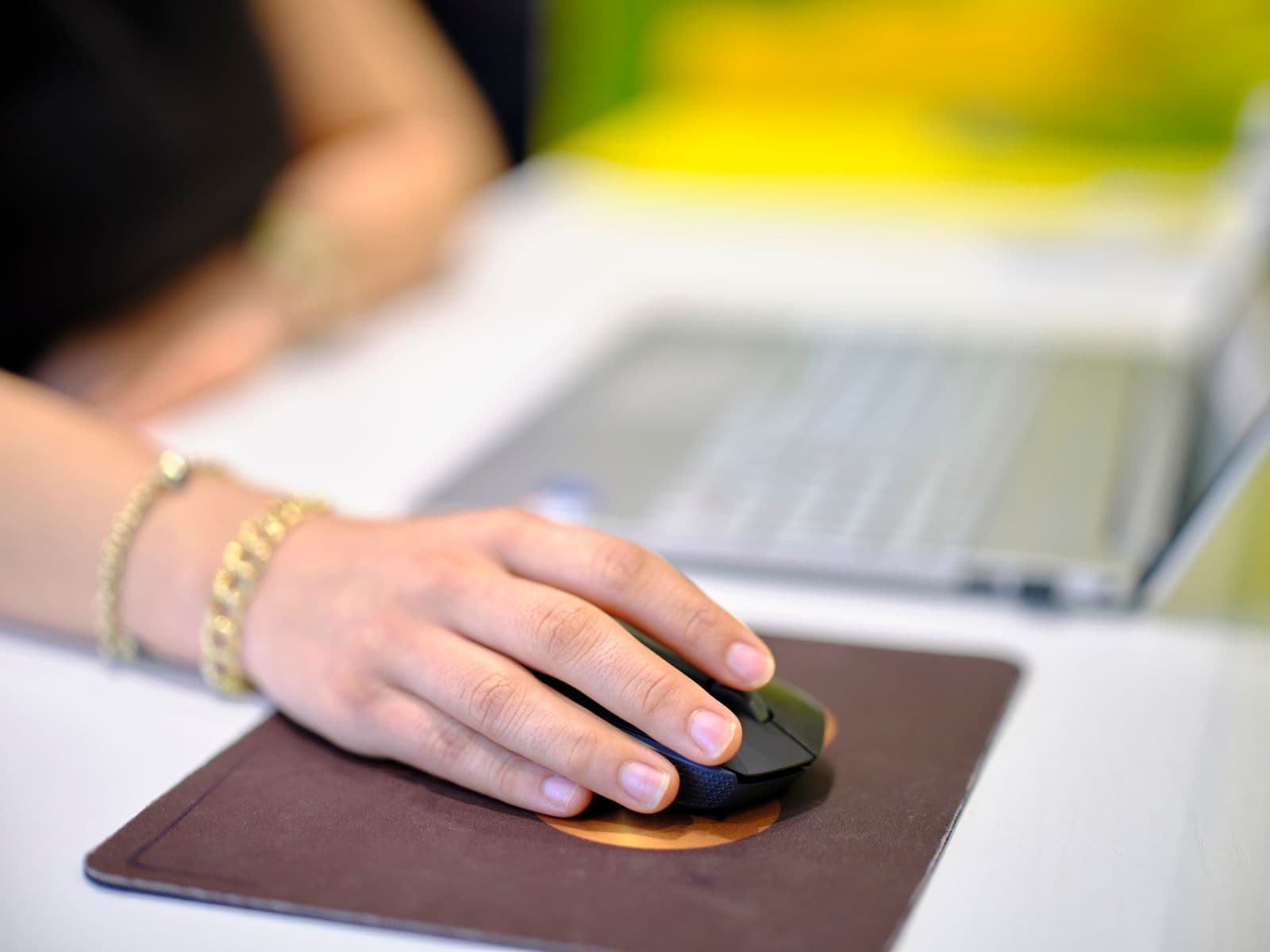 A hand resting palm-down on a black wireless mouse on a brown mouse pad, with a laptop out of focus behind it
