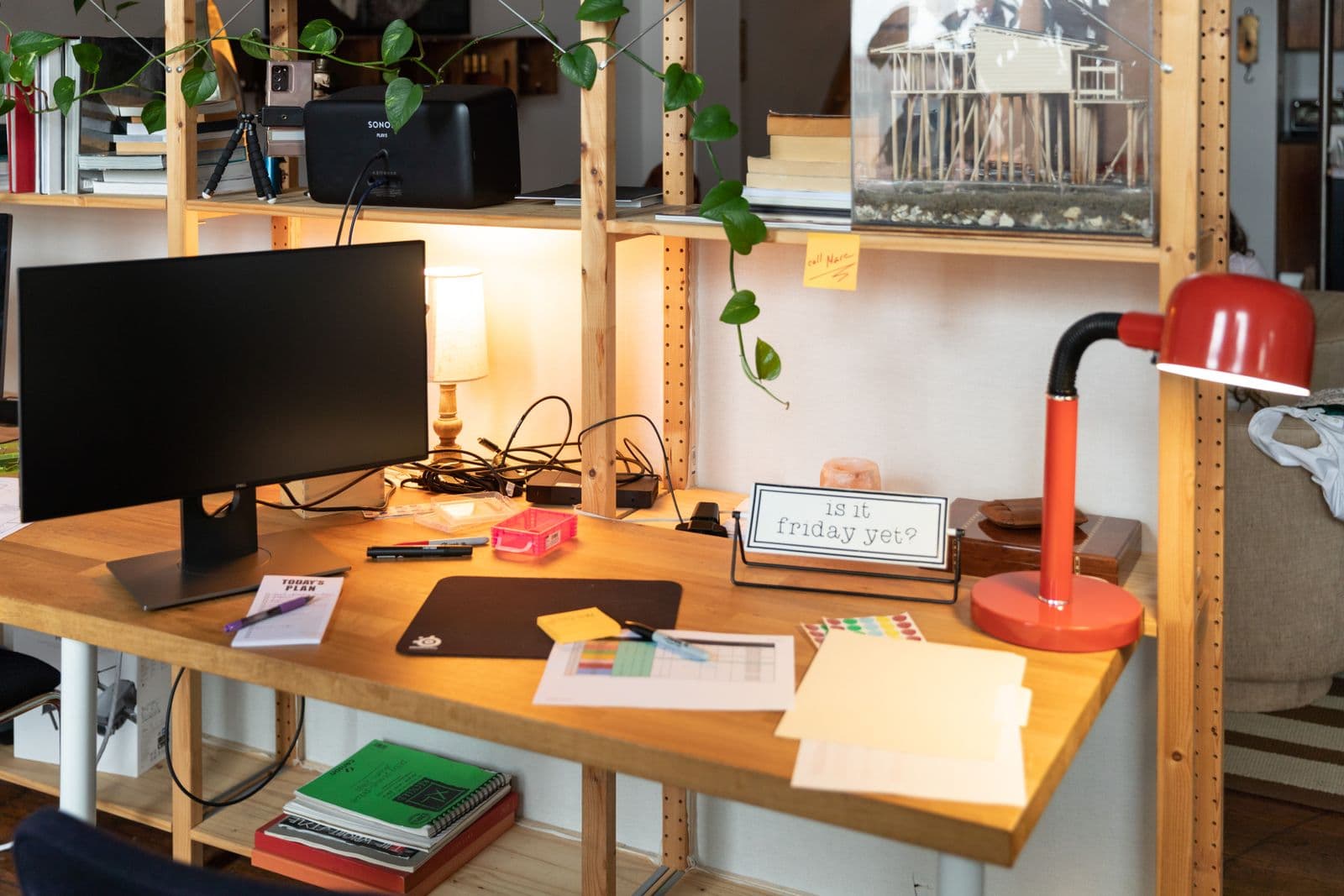 A wooden desk with a monitor, a red desk lamp, plants and office supplies