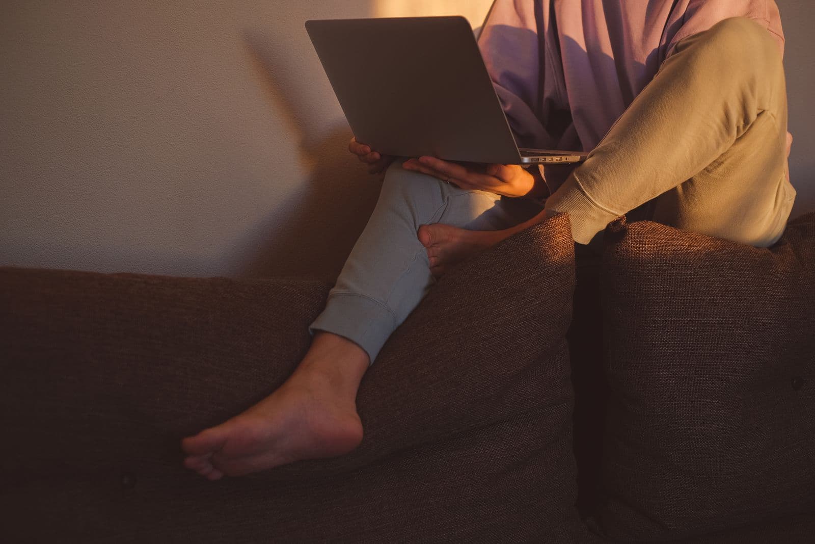 A person sitting barefoot and slouched on a couch with a laptop balanced on one knee