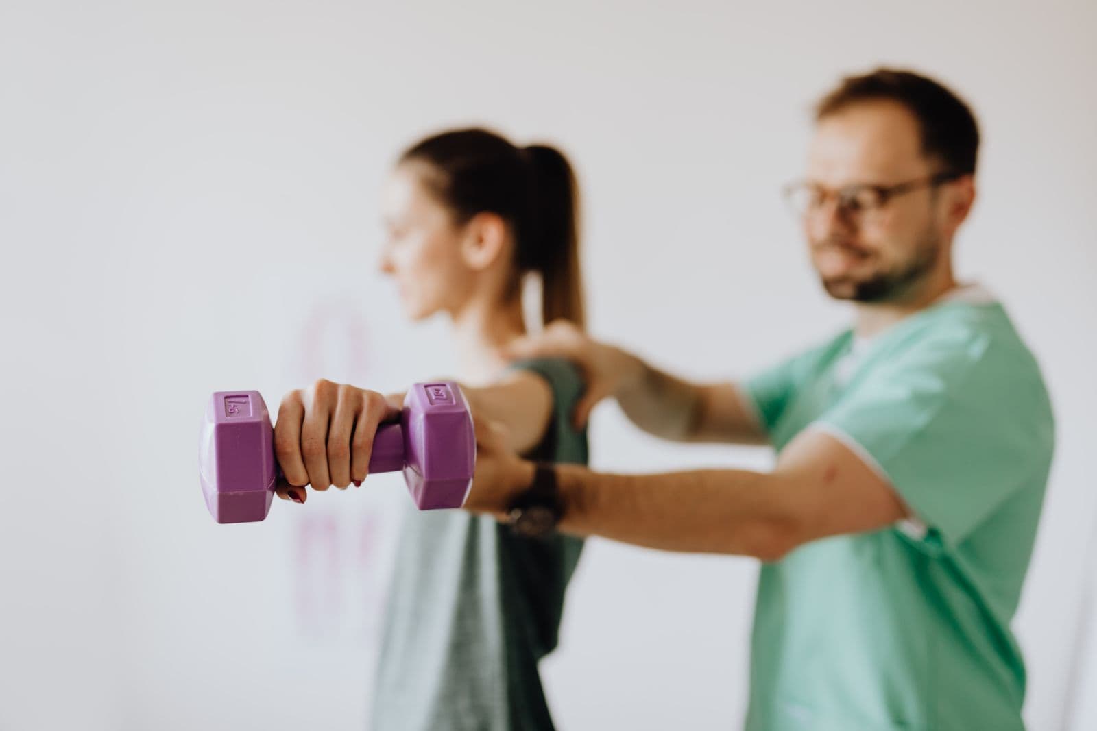 A woman holds a light purple dumbbell at arm's length while a physiotherapist steadies her shoulder