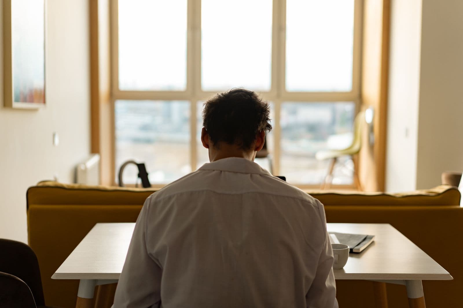 Back view of a man sitting at an office desk in a white shirt, in front of a window