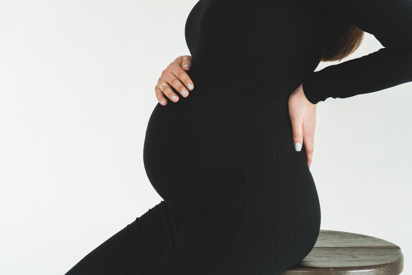 A pregnant woman in a black ribbed dress sitting on a wooden stool with one hand on her bump and the other pressed to her lower back