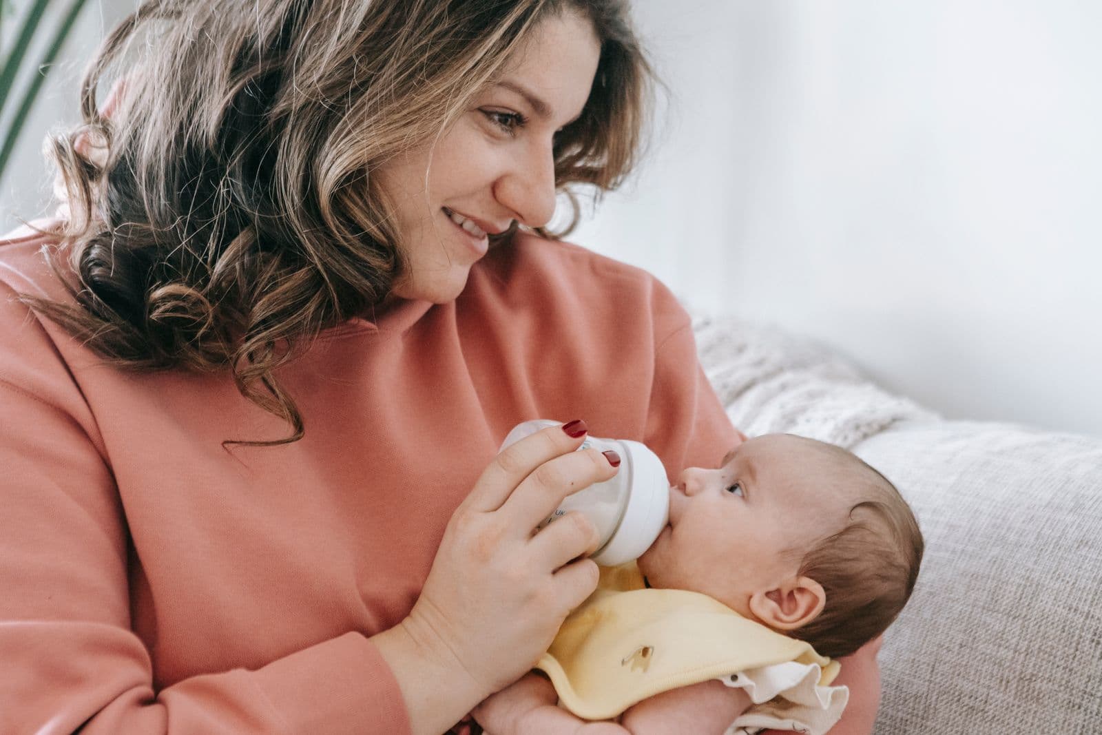 A mother sitting on a couch with shoulders rounded forward while feeding her infant