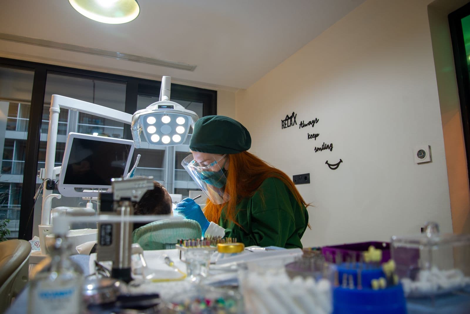 A dentist in green scrubs and a face shield leaning in over a patient while seated in a treatment room