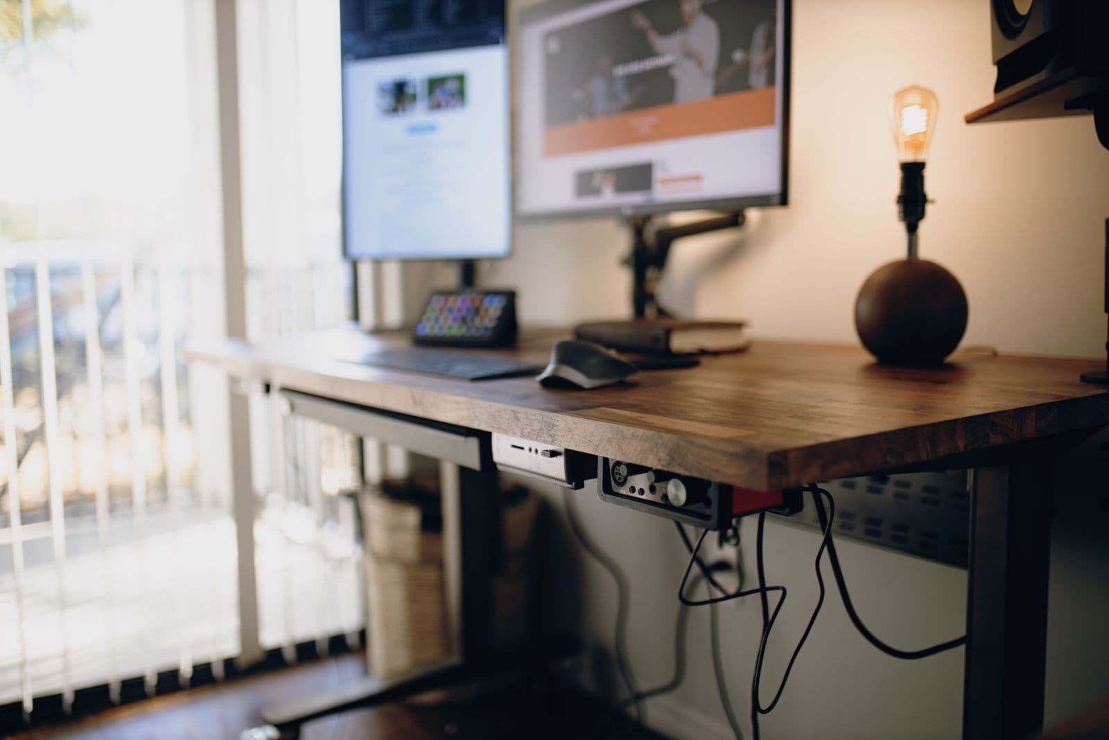 A height-adjustable desk with a wooden top, cable tray and monitor by a window