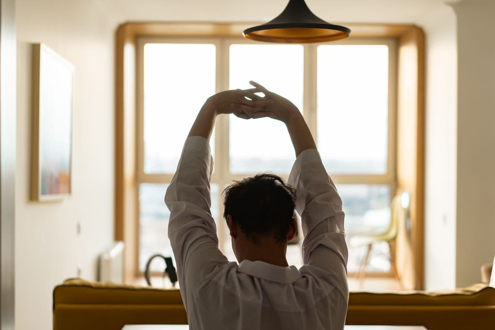 A person stretching both arms overhead in front of a sunlit window