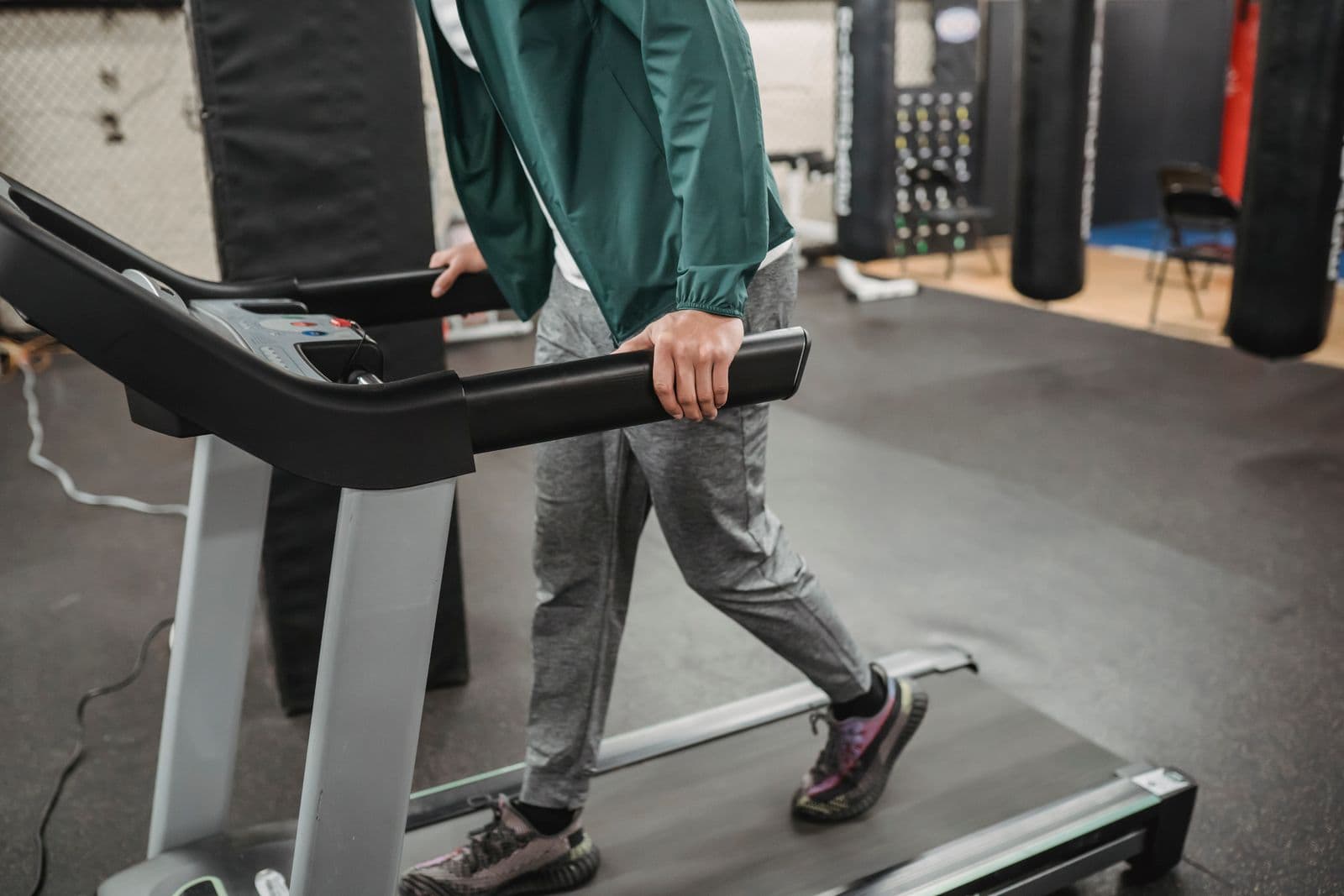 A person in grey joggers and a green jacket walking slowly on a treadmill with both hands resting on the side rails
