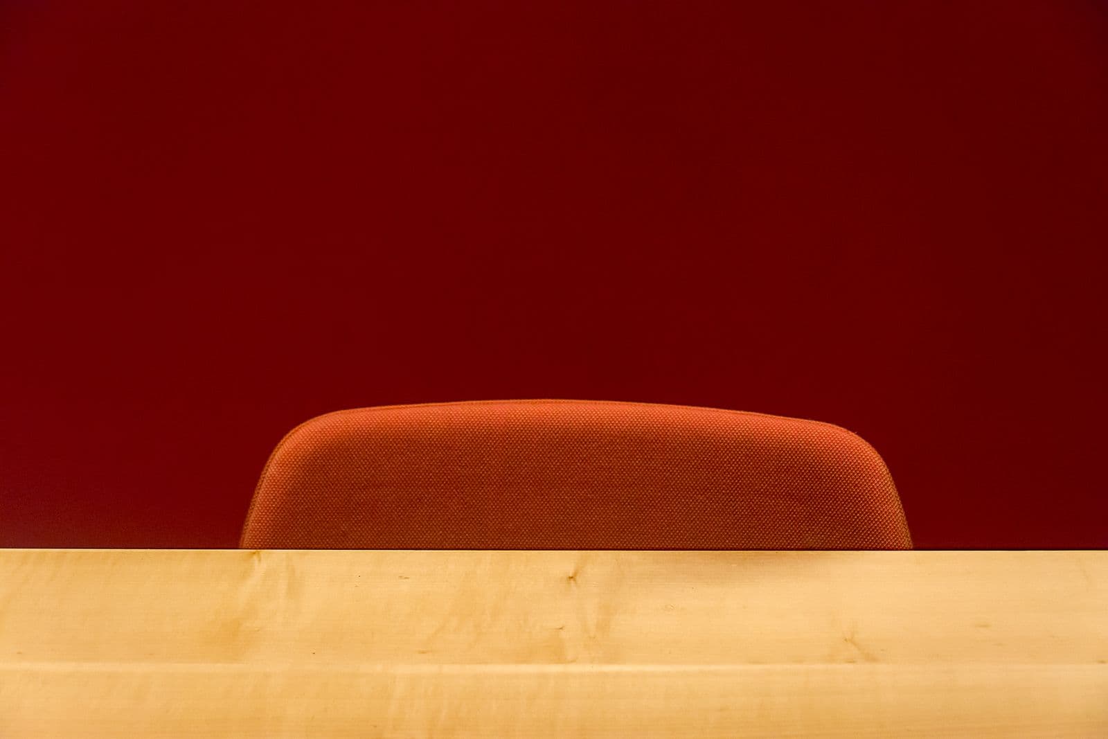 The upholstered back of an orange office chair behind a pale wooden desk, against a dark red wall