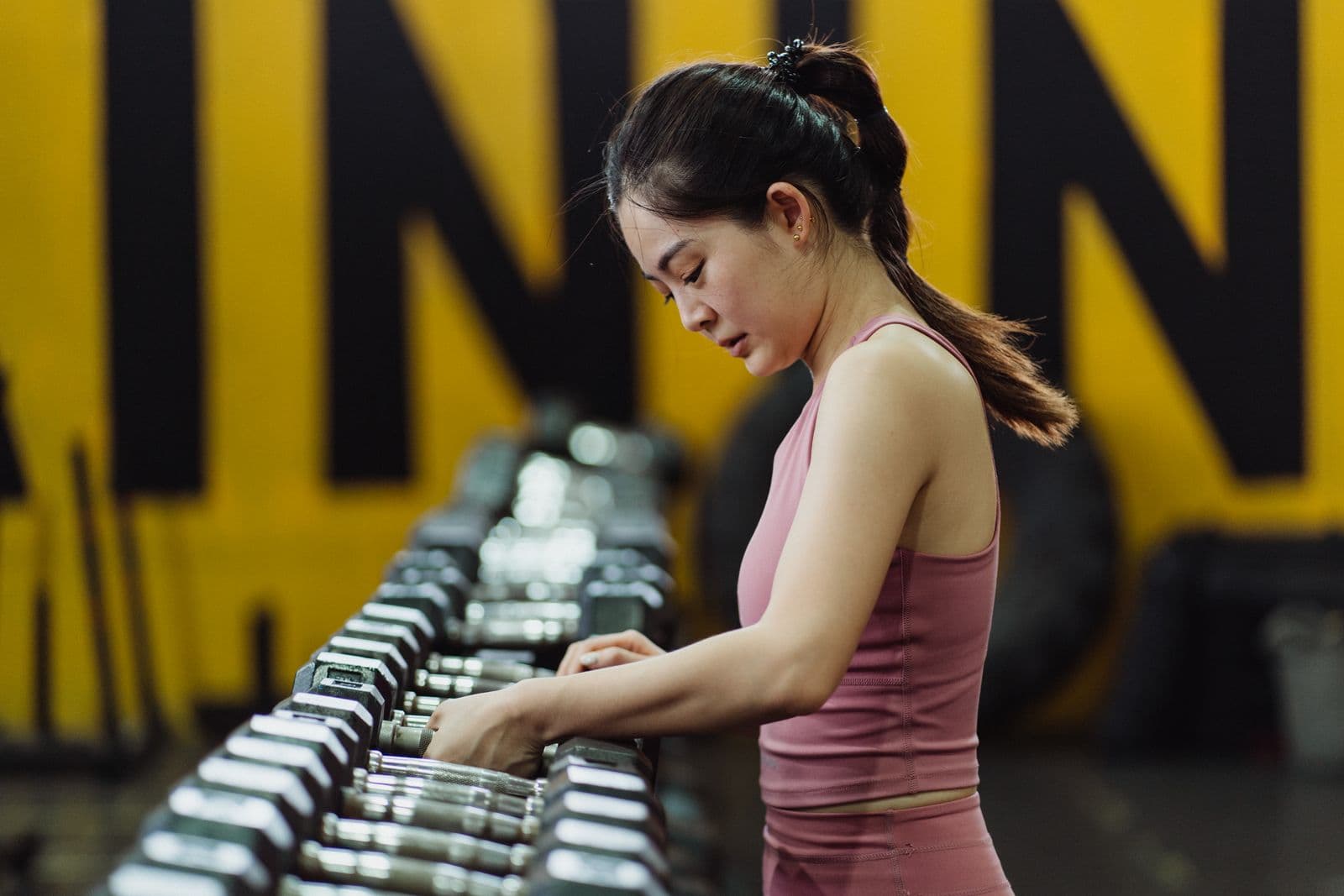 A woman in a pink training top reaches for a hex dumbbell on a long gym rack