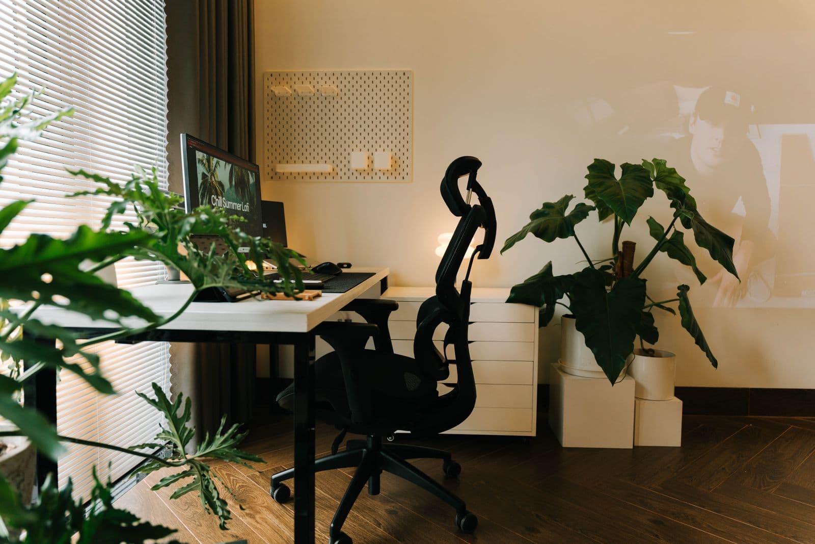 A black mesh office chair with a headrest pulled up to a white desk in a home office with plants